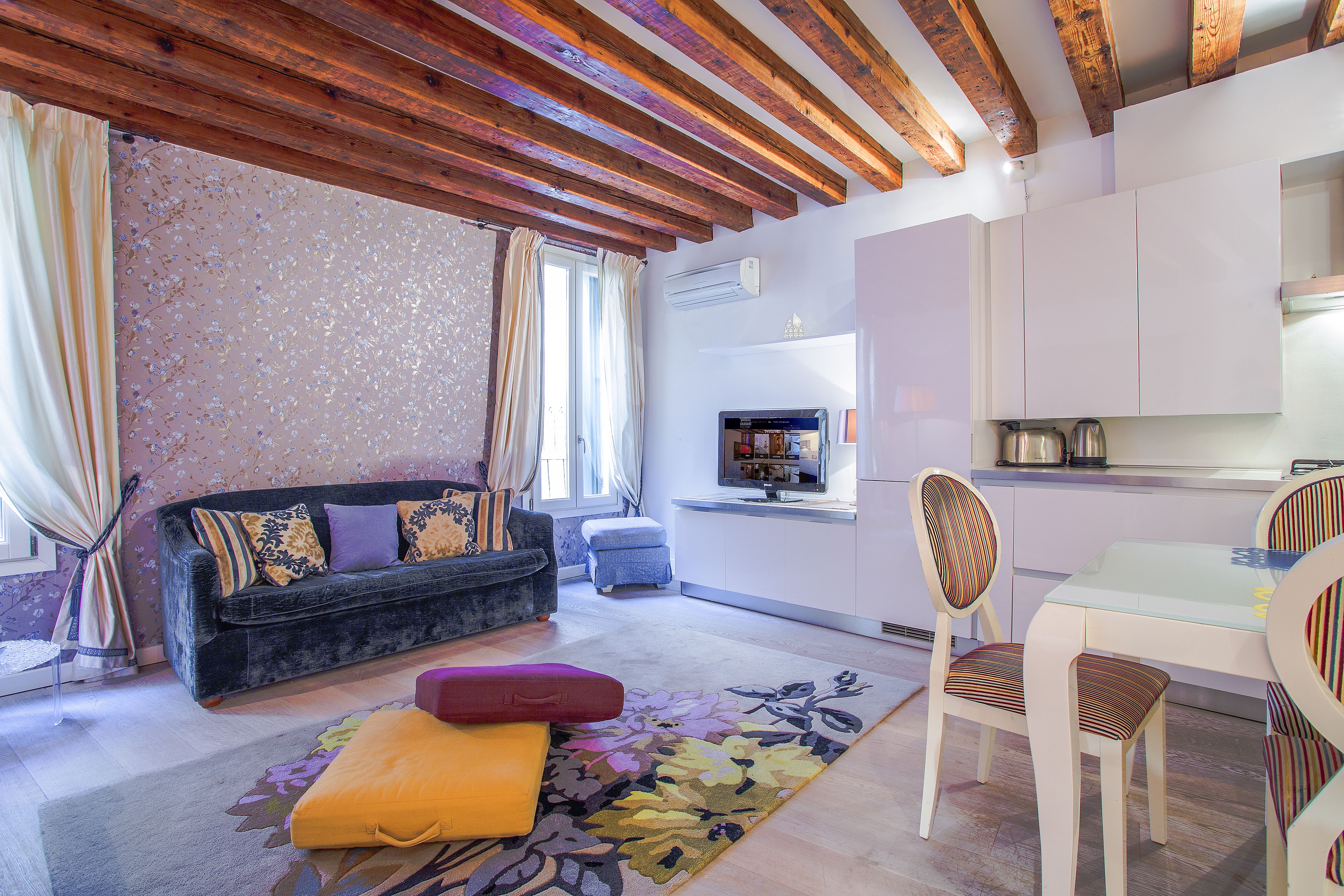 Sala de estar con sofá, reposapiés y sillas, cocina con televisor y techo de madera.