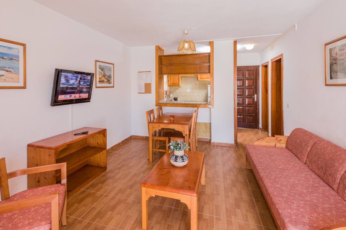 A living room with a wooden dining table, chairs, a sofa, a TV, and a kitchen area with wooden cabinets and a hanging light fixture.