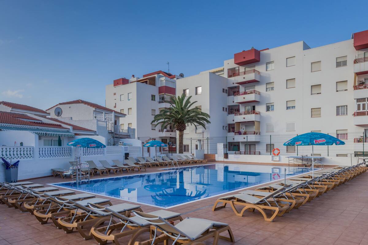 A swimming pool with lounge chairs and umbrellas, surrounded by multi-story buildings with balconies.