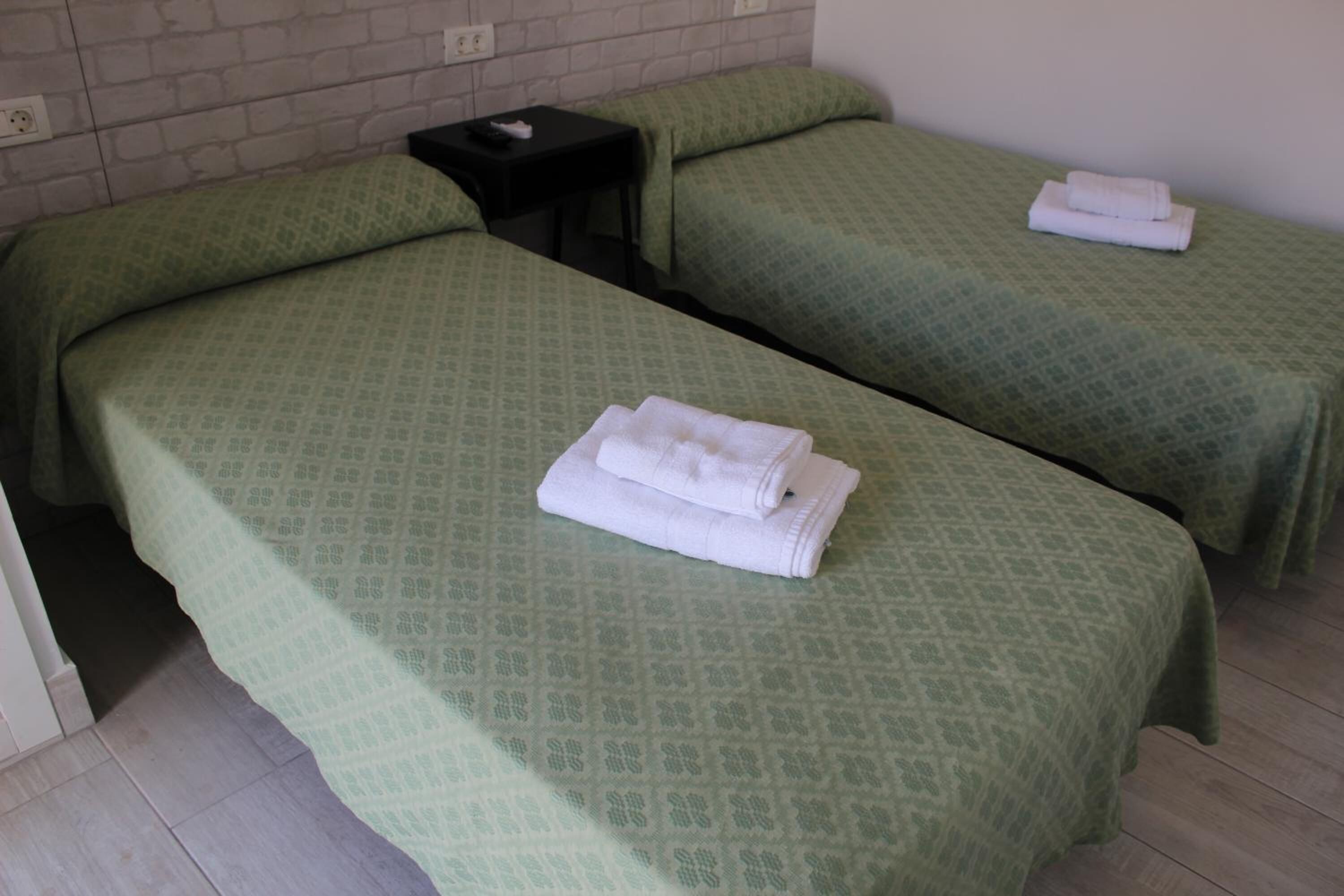 A room with two beds, each with green patterned bedding and a folded white towel on the bed. There is a small black nightstand with a remote control on it.