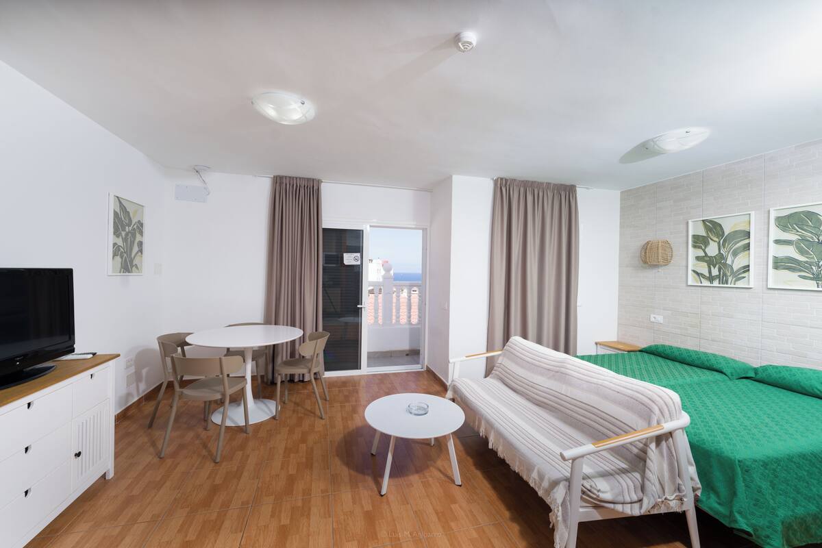A modern hotel room with a flat-screen TV, a dining table with chairs, a sofa, and a bed with a green cover.