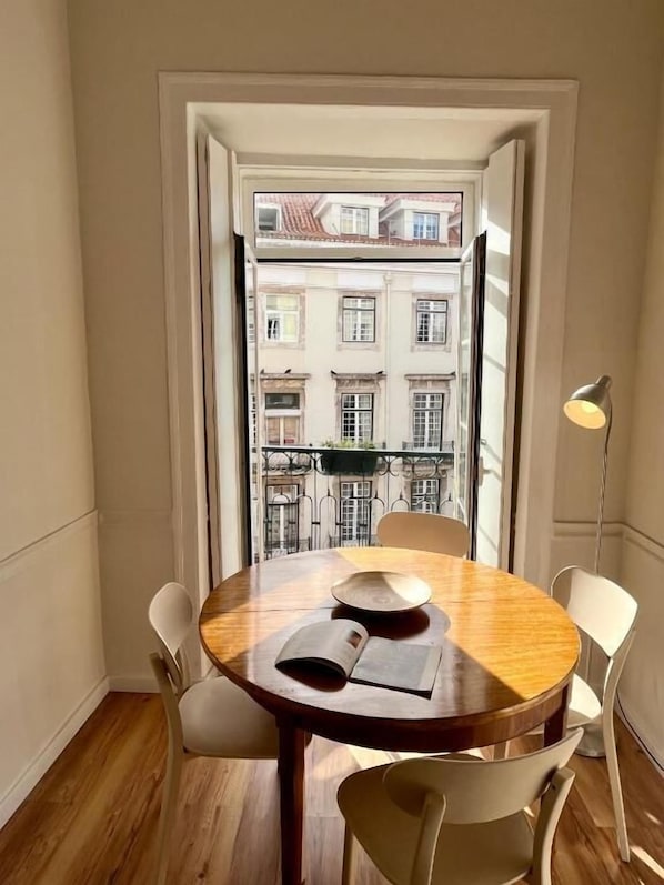 A small dining area with a round table and four chairs, a window with a view of buildings, and a floor lamp.