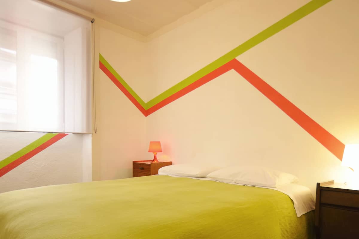 Een kamer met een bed, nachtkastje, lamp en raam.