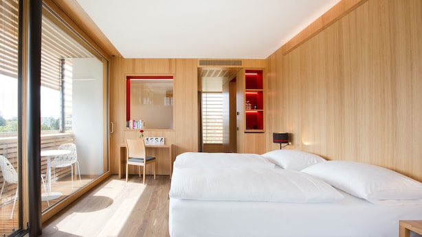 Habitació d'hotel moderna amb sòl de fusta, un llit gran amb roba de llit blanca, un escriptori amb una cadira i una porta de vidre que dona a una zona exterior.