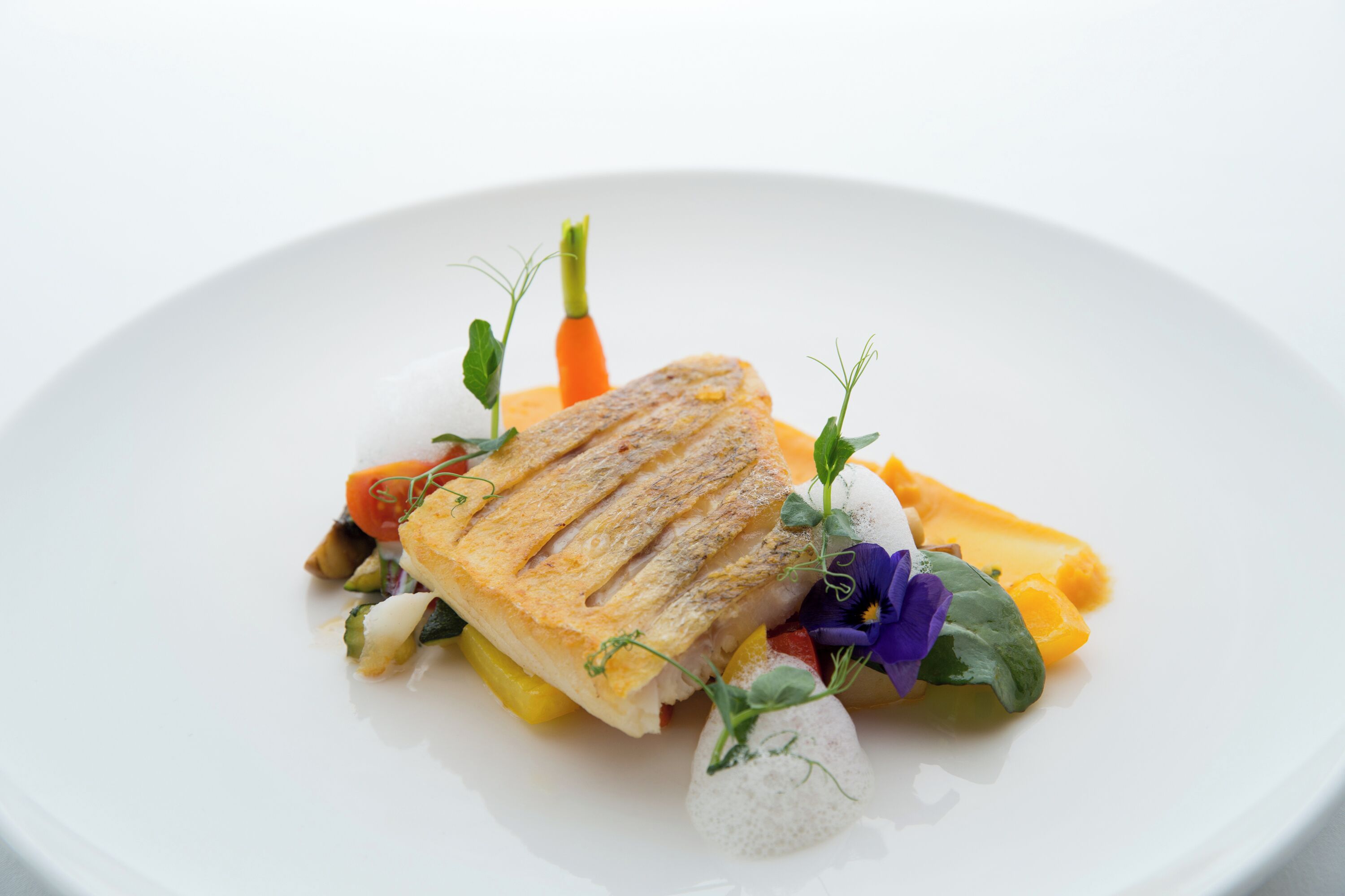 Un plato blanco con pescado a la parrilla, verduras y una flor decorativa.