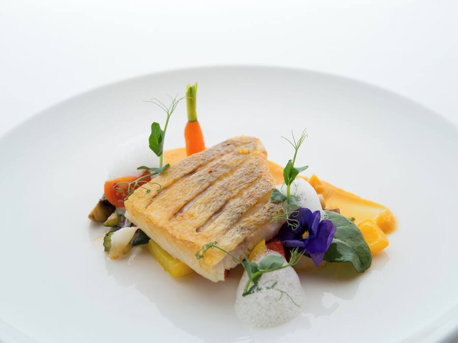 Un plato blanco con pescado a la parrilla, verduras y una flor decorativa.