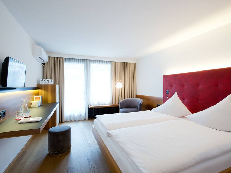 Habitación de hotel con dos camas, un escritorio con televisor, una silla, una mesita de noche con lámpara y una ventana con cortinas.