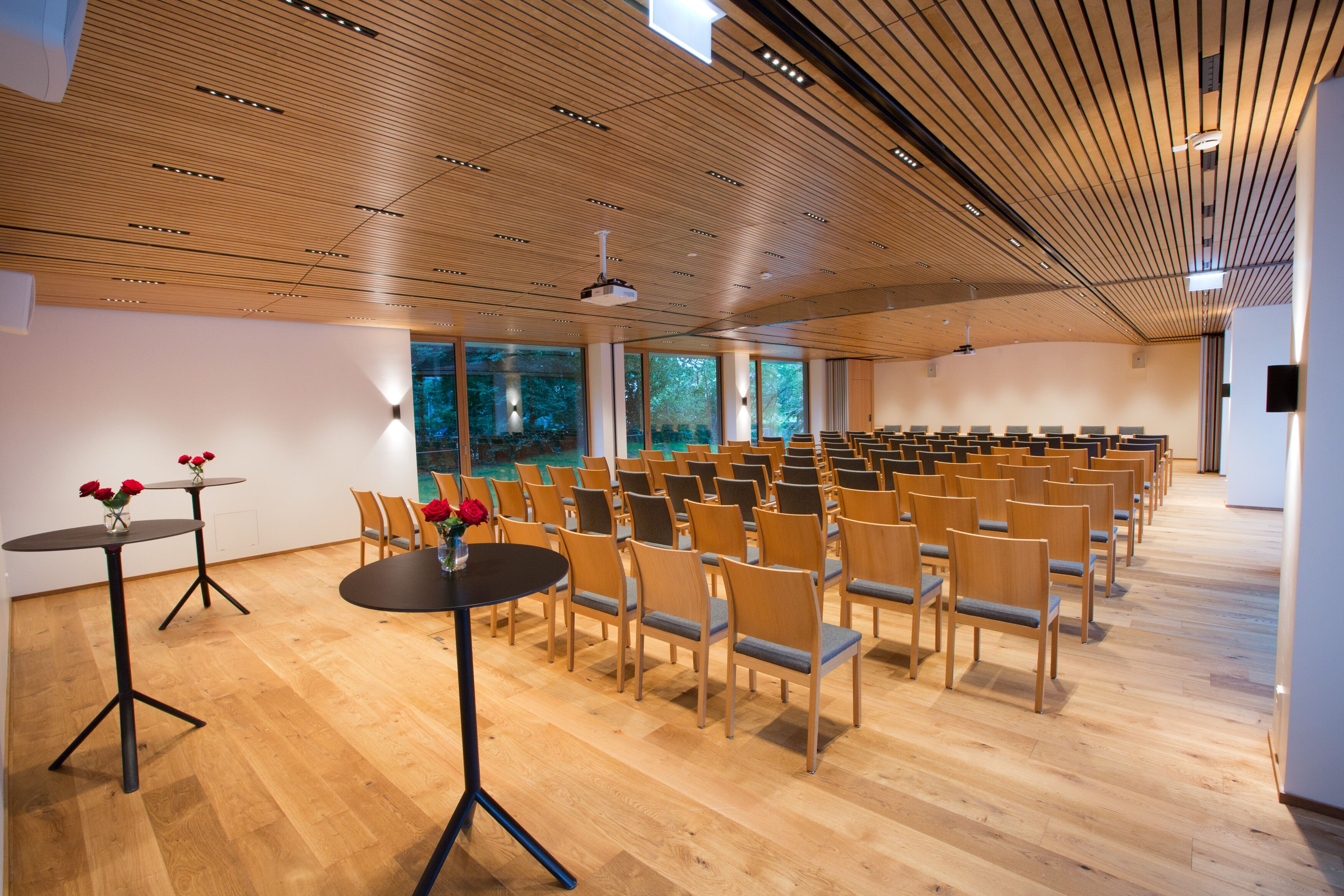 A conference room with rows of chairs, a round table with a vase of flowers, and large windows with a view of trees.