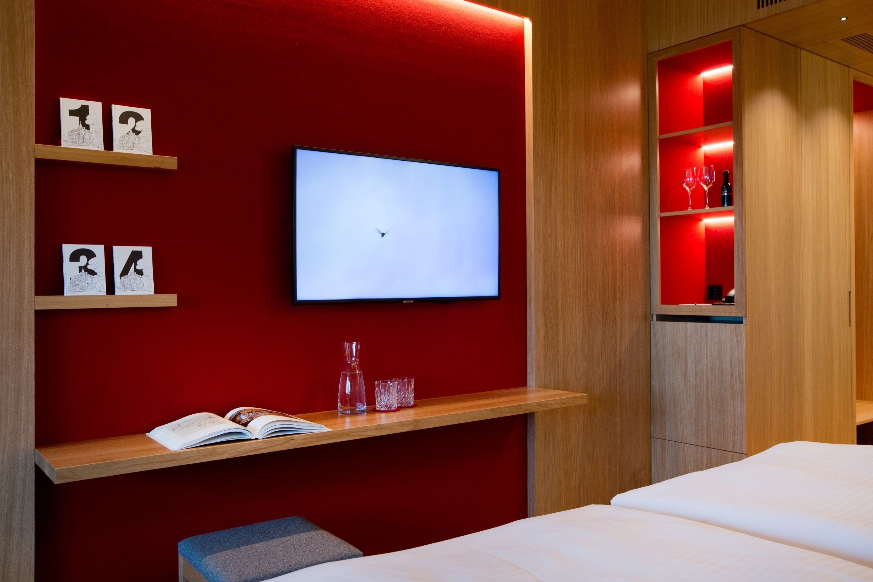 Una habitación de hotel moderna con una pared de acento roja, un televisor montado, estantes de madera con libros y vasos, y una cama con ropa blanca.