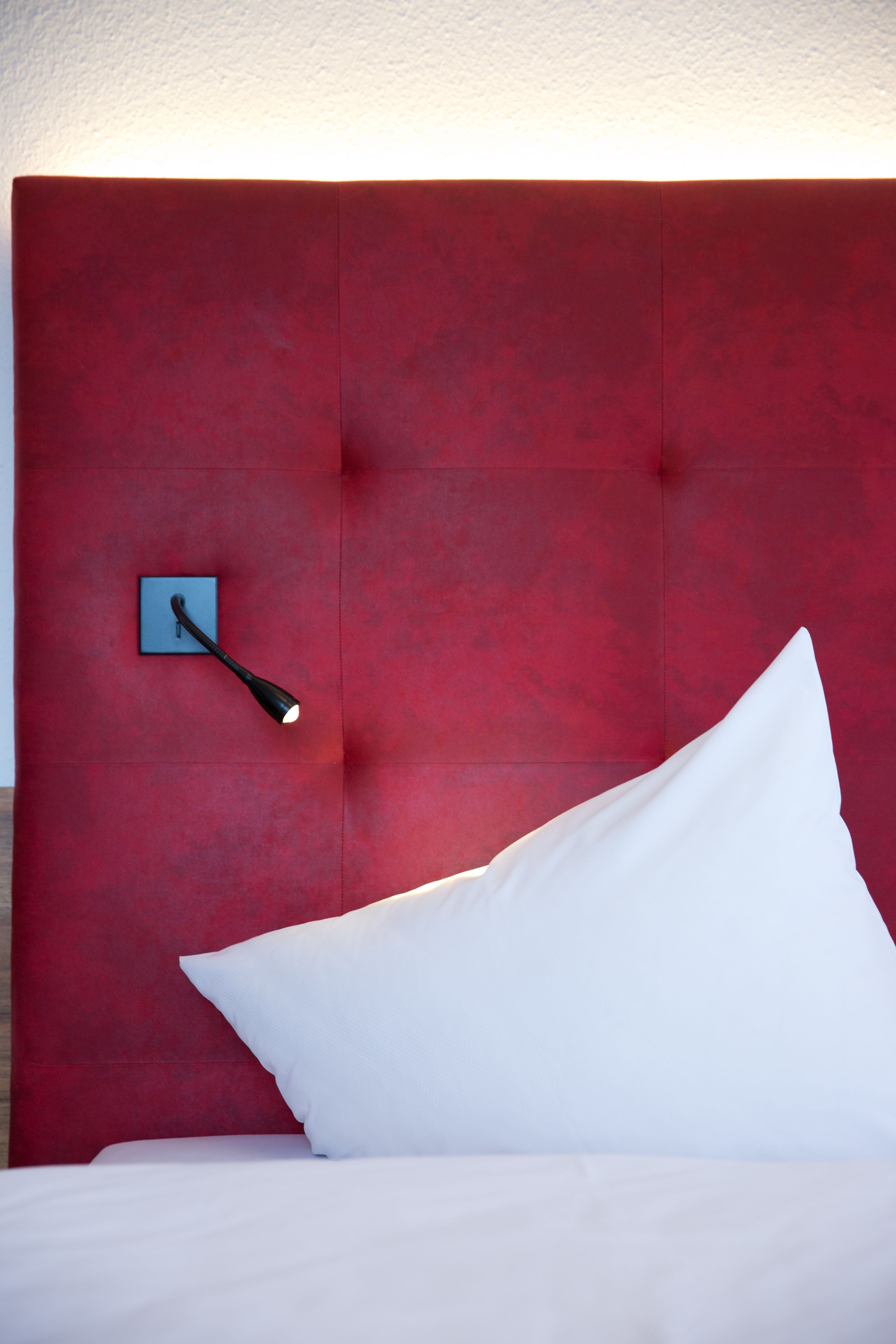 A bed with white bedding, a red upholstered headboard, and a wall-mounted light fixture.