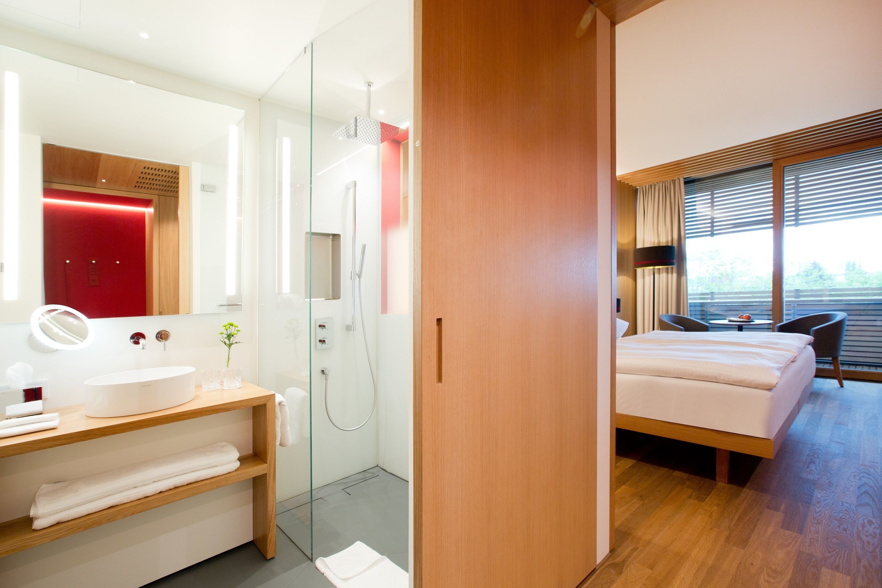 Habitación de hotel moderna con suelo de madera, cama grande, mesita de noche, espejo, zona de ducha y vistas al exterior a través de la ventana.