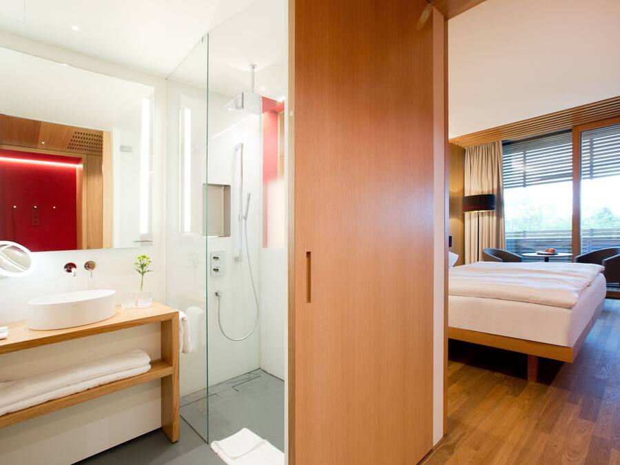 Habitación de hotel moderna con suelo de madera, cama grande, mesita de noche, espejo, zona de ducha y vistas al exterior a través de la ventana.