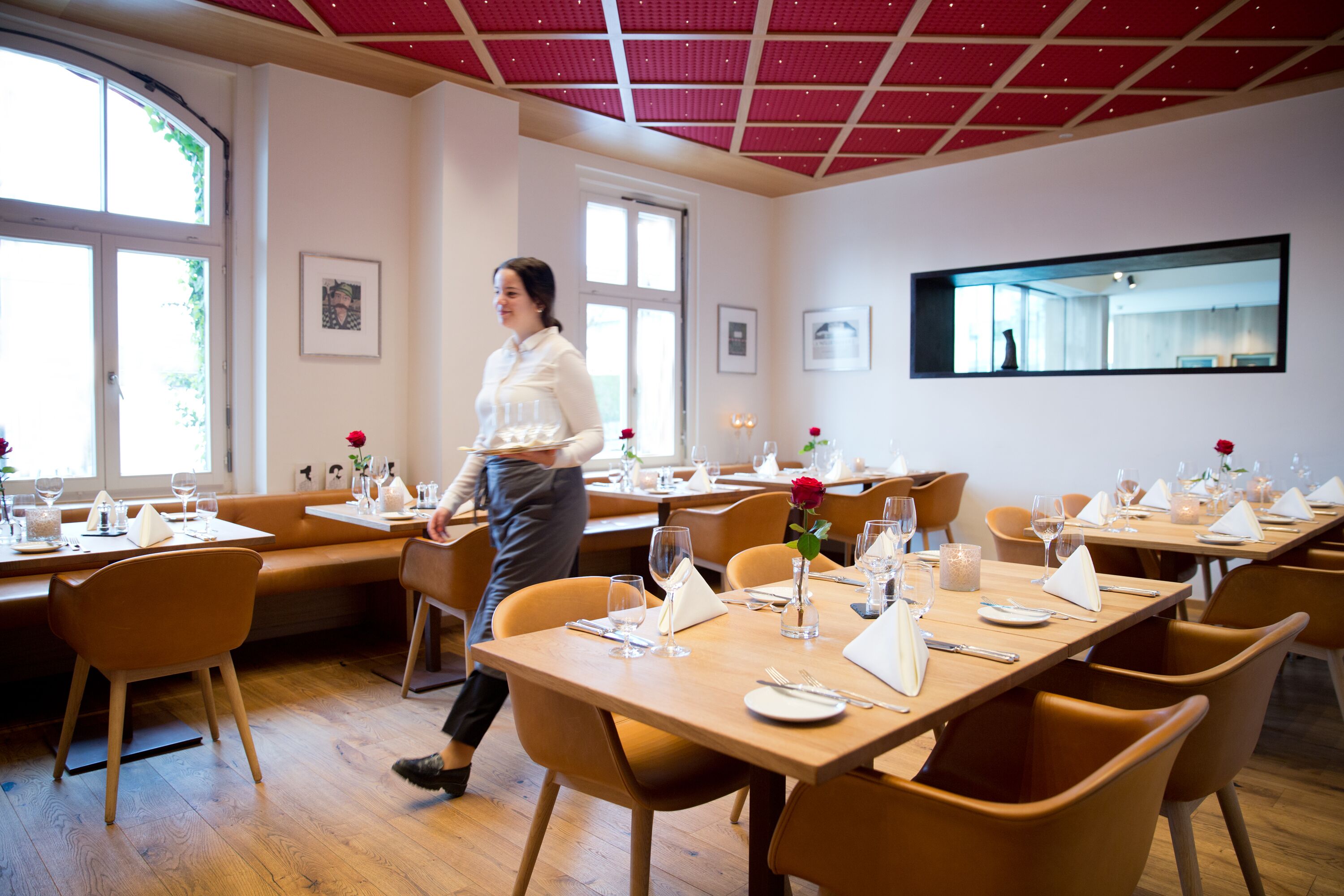 Un restaurante con mesas y sillas de madera, techo rojo y cuadros en las paredes.