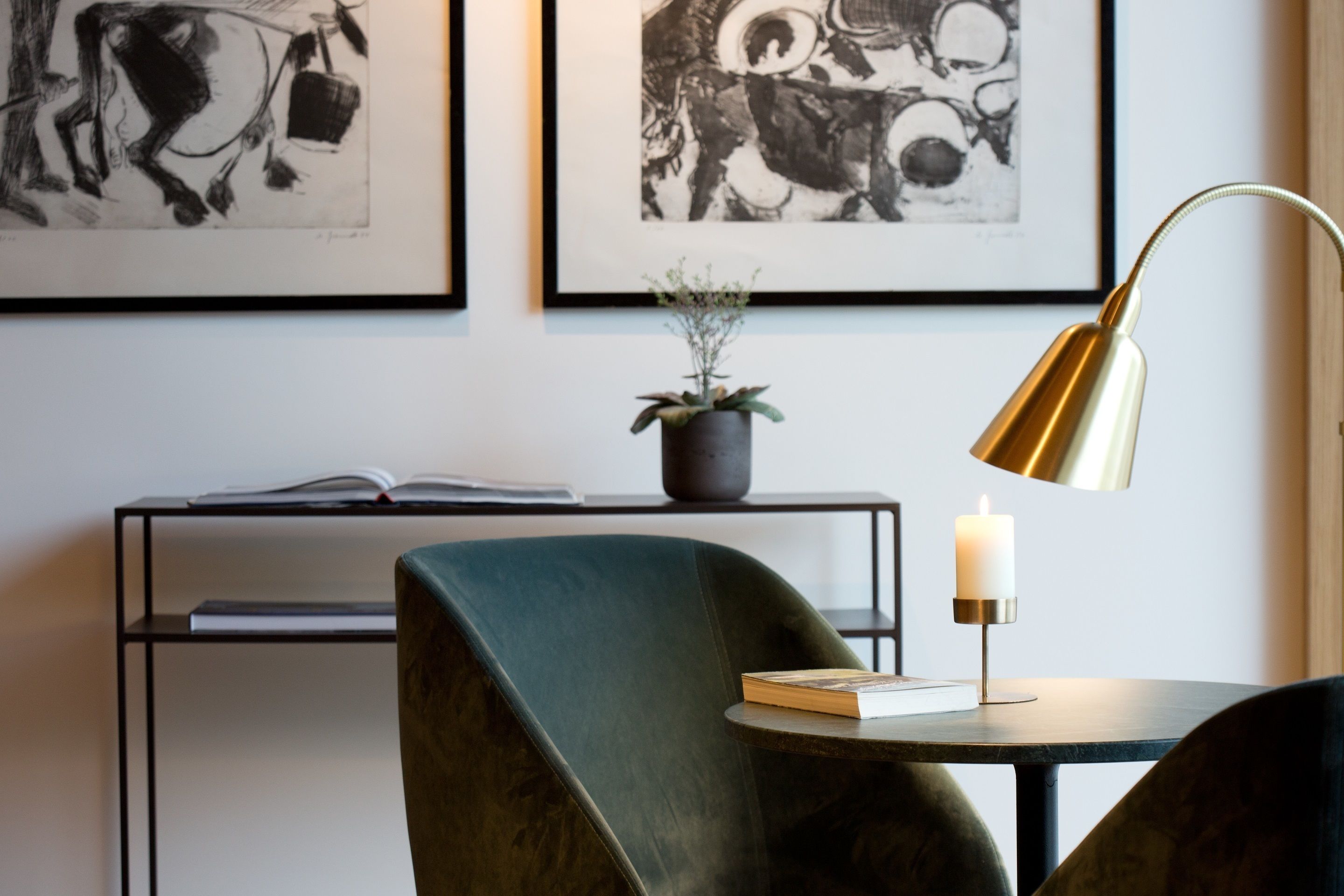 A modern room with a green armchair, a round table with a book and a candle, a floor lamp, and two framed abstract paintings on the wall.