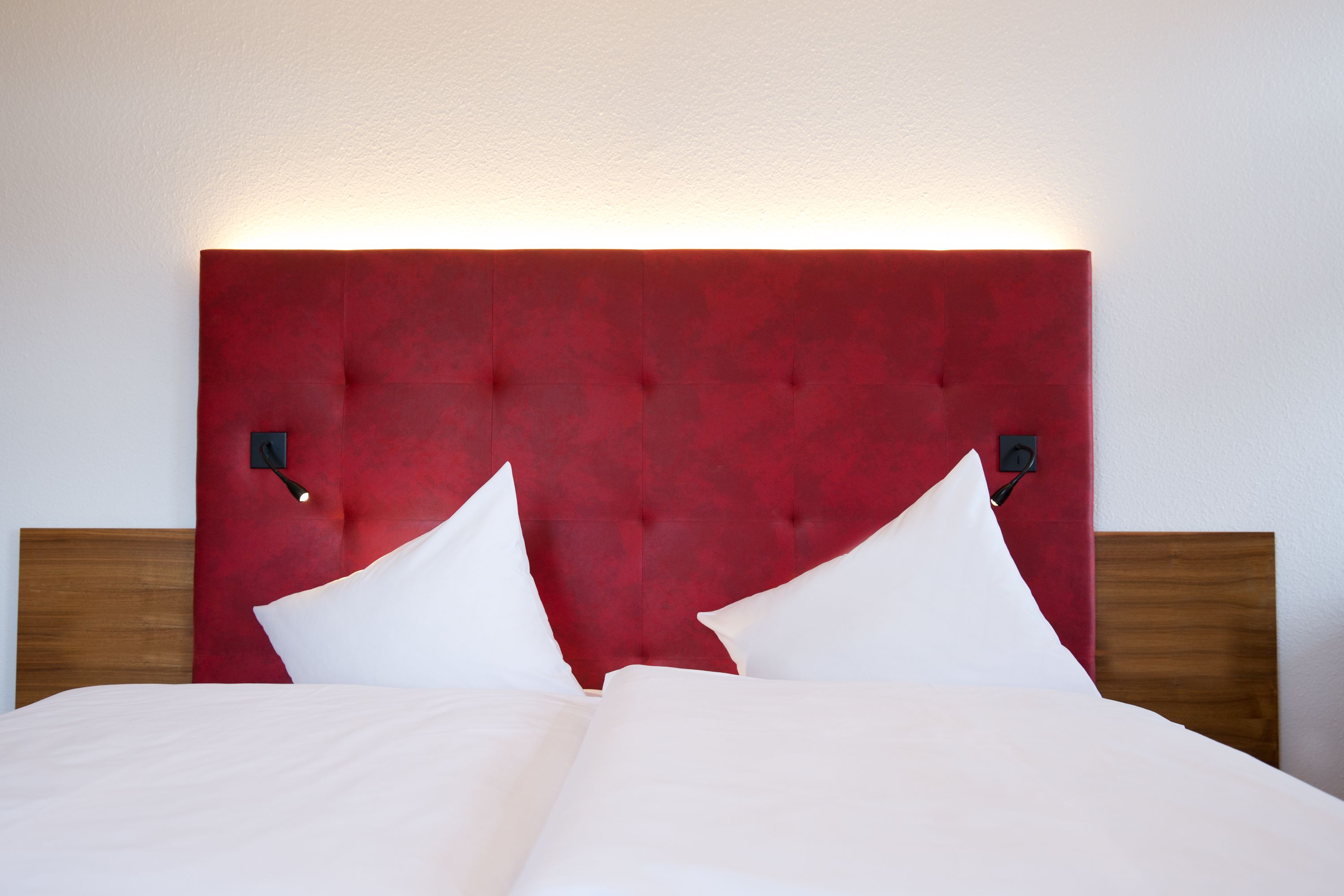A hotel room with a red upholstered headboard, two white pillows, and a wooden headboard on the left.