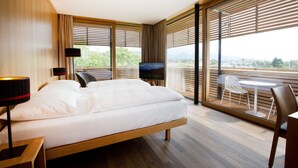 Habitació d'hotel moderna amb un llit gran, televisió, escriptori amb làmpada, cadira i un balcó amb taula i cadires.