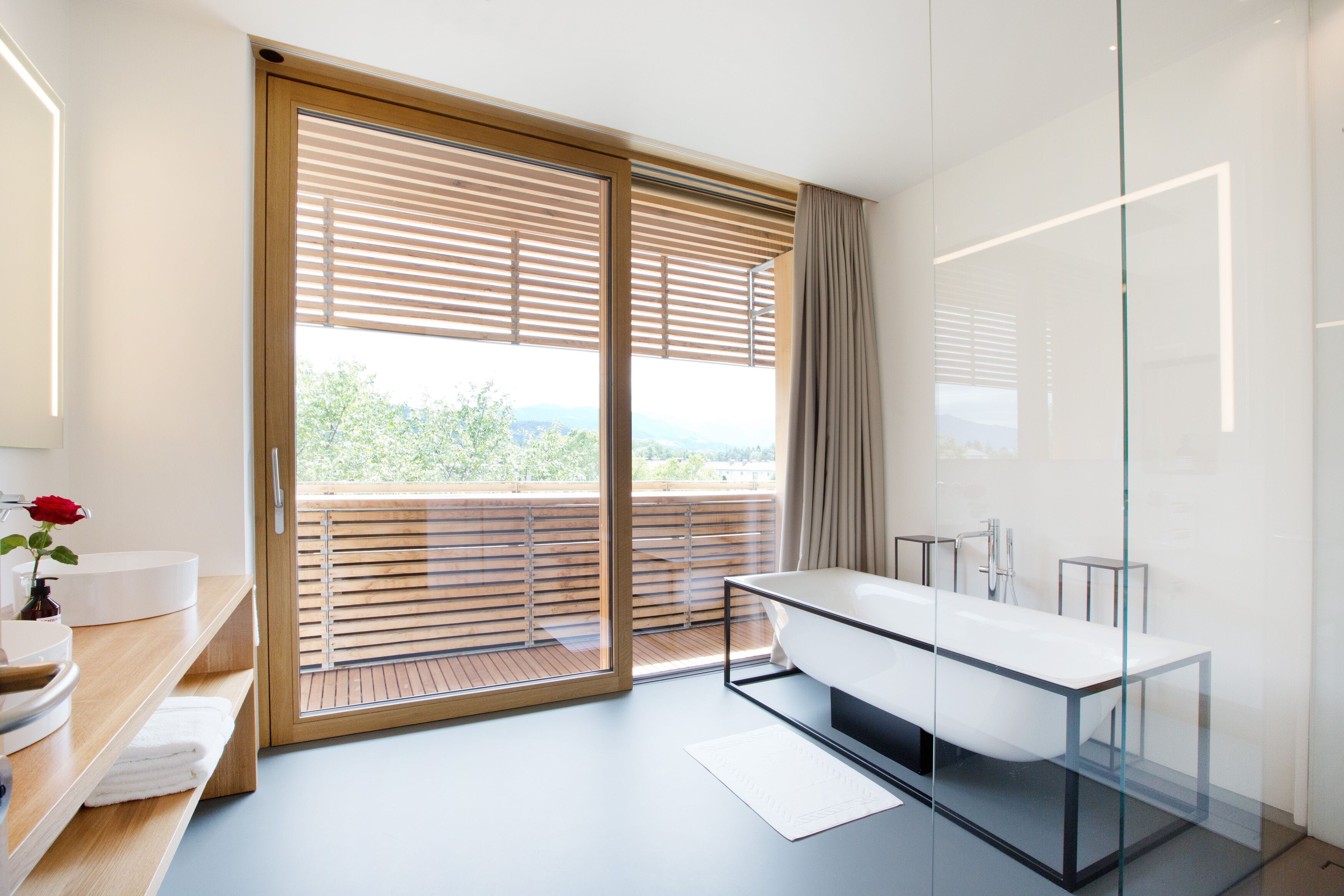 A modern bathroom with a glass-enclosed shower, a large window with wooden blinds, and a view of greenery outside.