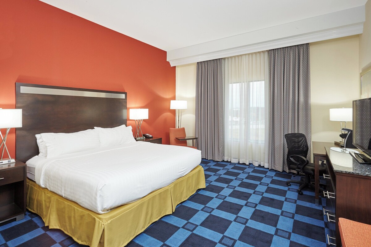 Une chambre d’hôtel avec un grand lit, deux tables de chevet avec des lampes, un bureau avec une chaise et une grande fenêtre avec des rideaux.