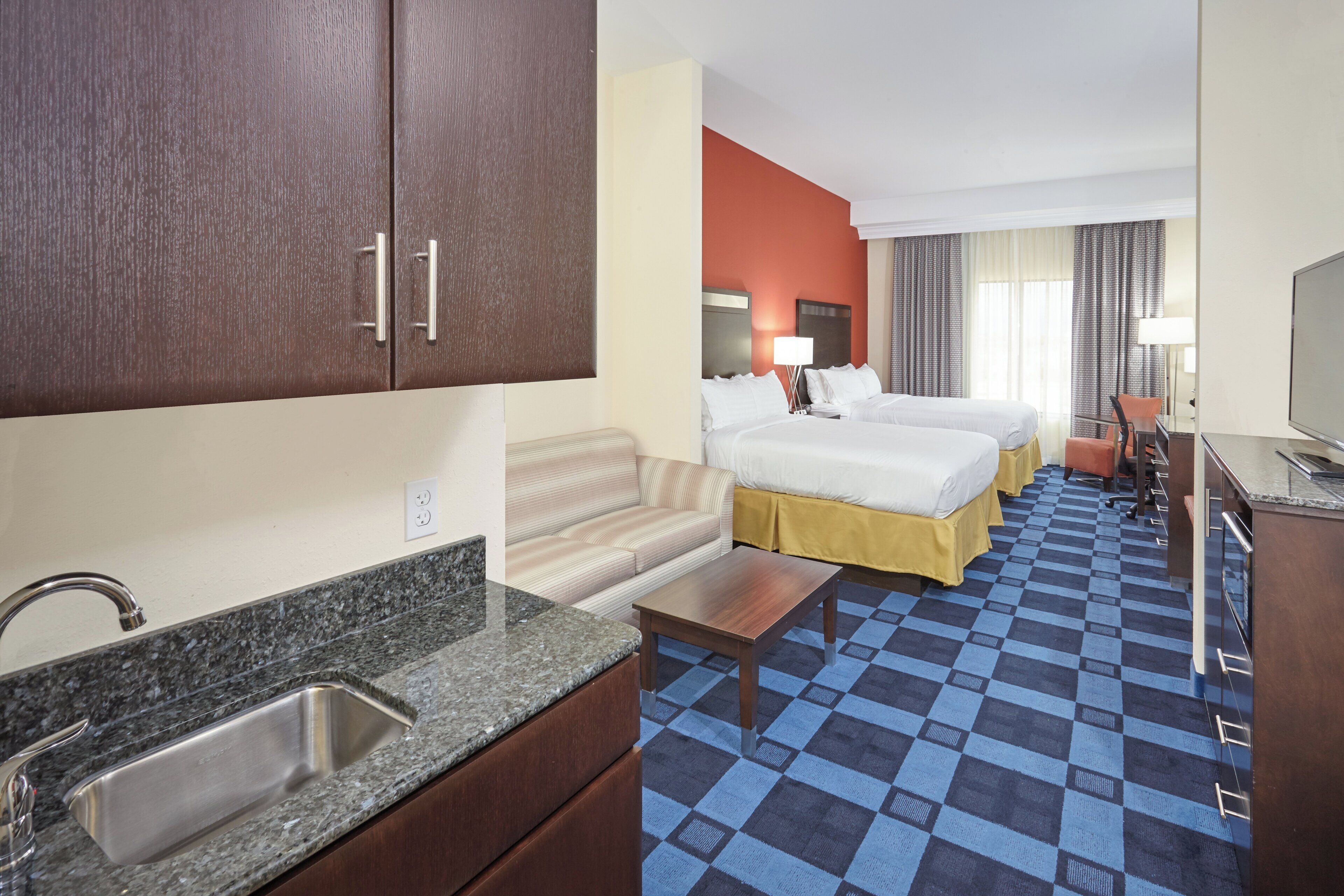 Quarto de hotel com cozinha compacta, duas camas, um sofá, uma TV e piso xadrez.