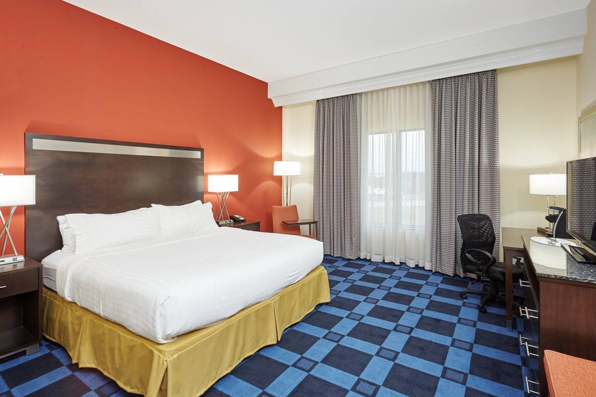 Une chambre d’hôtel avec un grand lit, deux tables de chevet avec des lampes, un bureau avec une chaise et une grande fenêtre avec des rideaux.