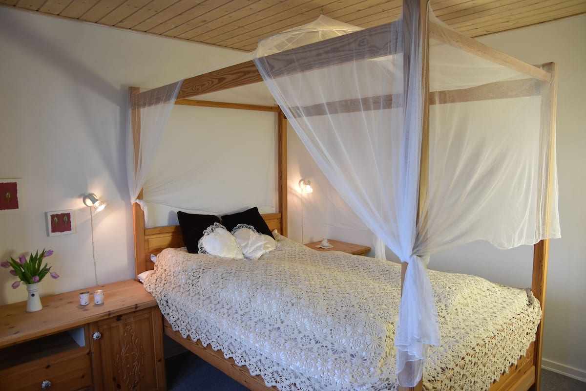 Ein Bett mit Baldachin, ein Nachttisch aus Holz mit Blumenvase und eine Wandleuchte.
