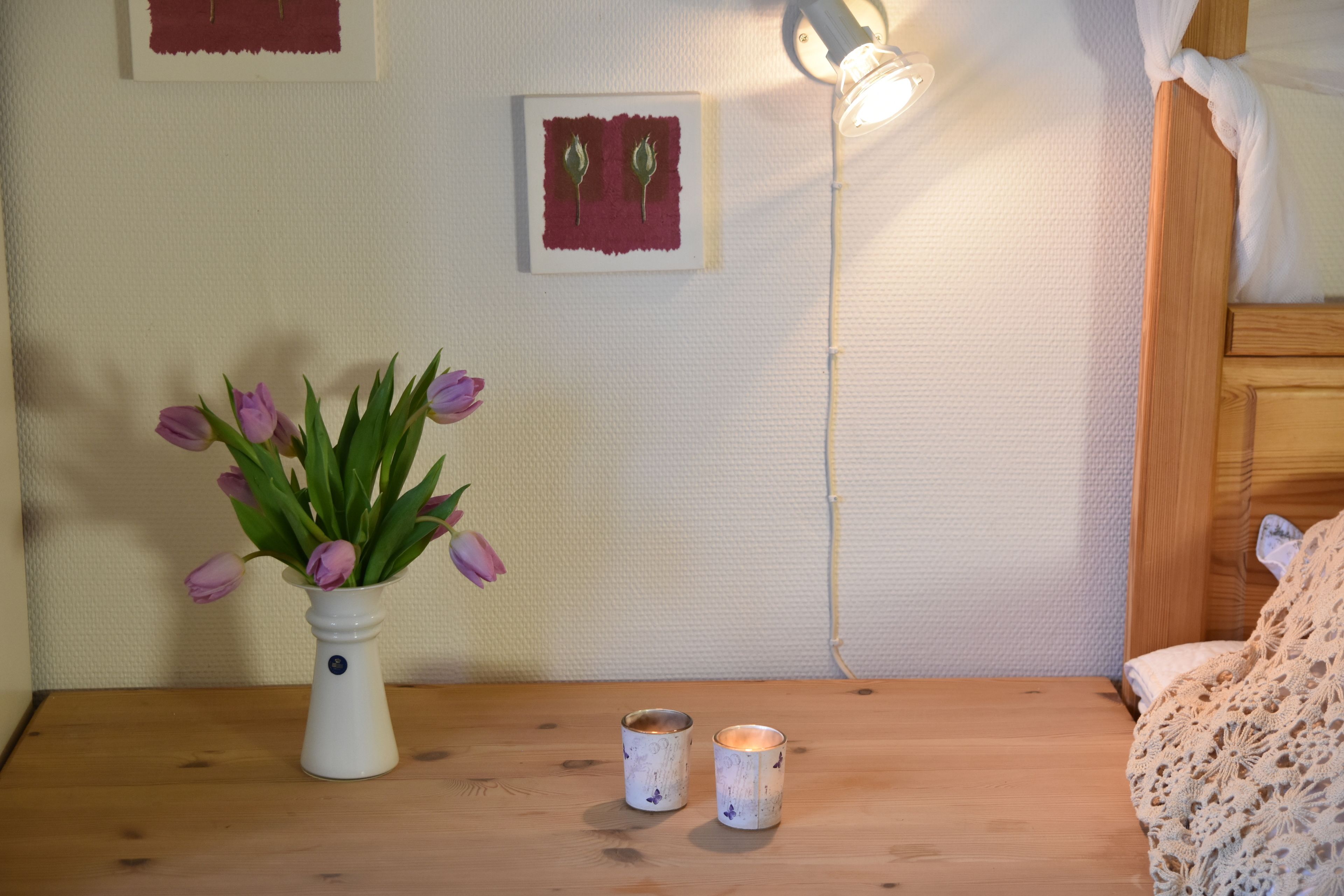 Una mesa de madera con un jarrón blanco de tulipanes morados, dos velas pequeñas y un cuadro en la pared.