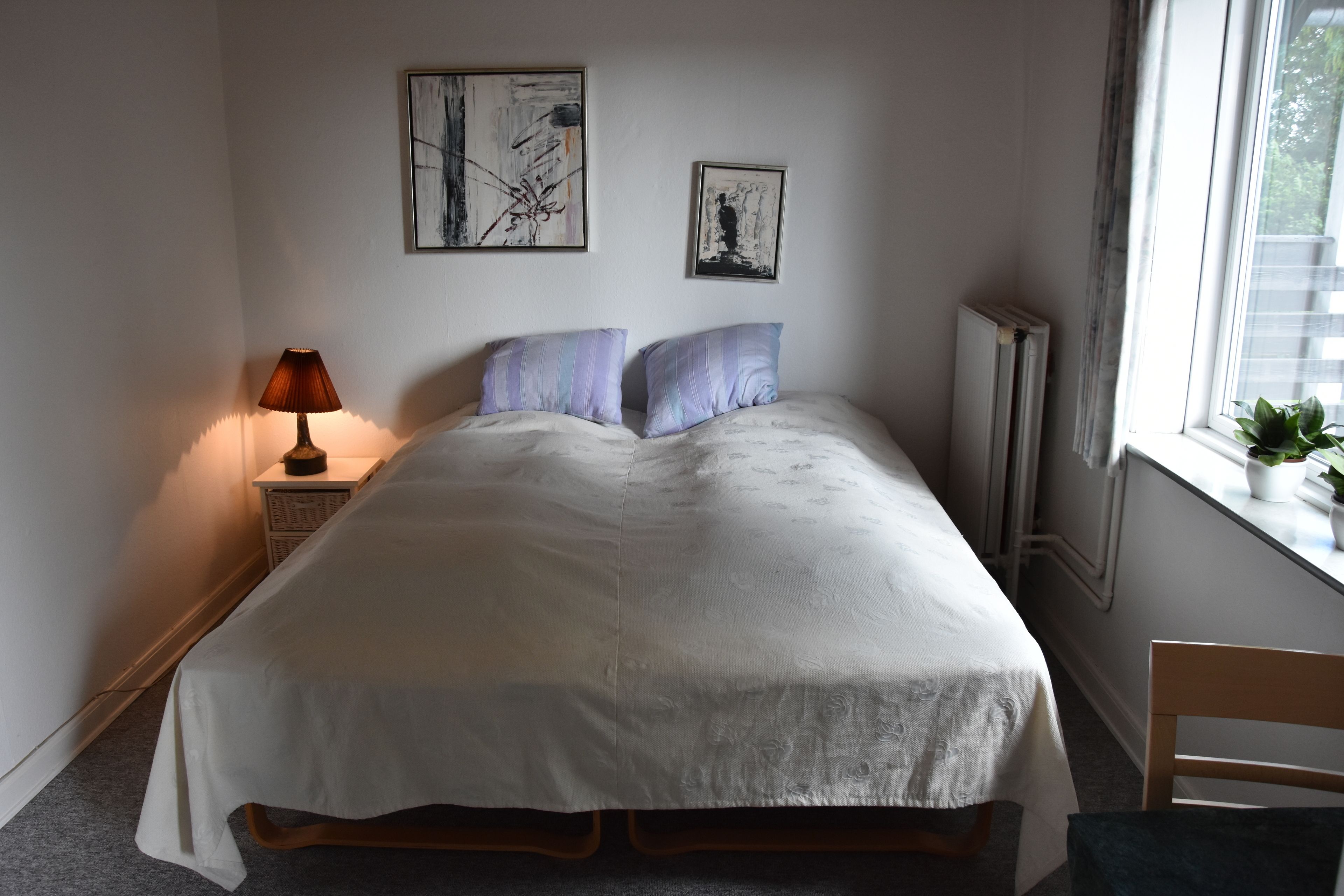 A neatly made bed with a white duvet, two pillows, a bedside lamp, and two framed artworks on the wall.