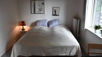 Una cama bien hecha con edredón blanco, dos almohadas, una lámpara de noche y dos obras de arte enmarcadas en la pared.