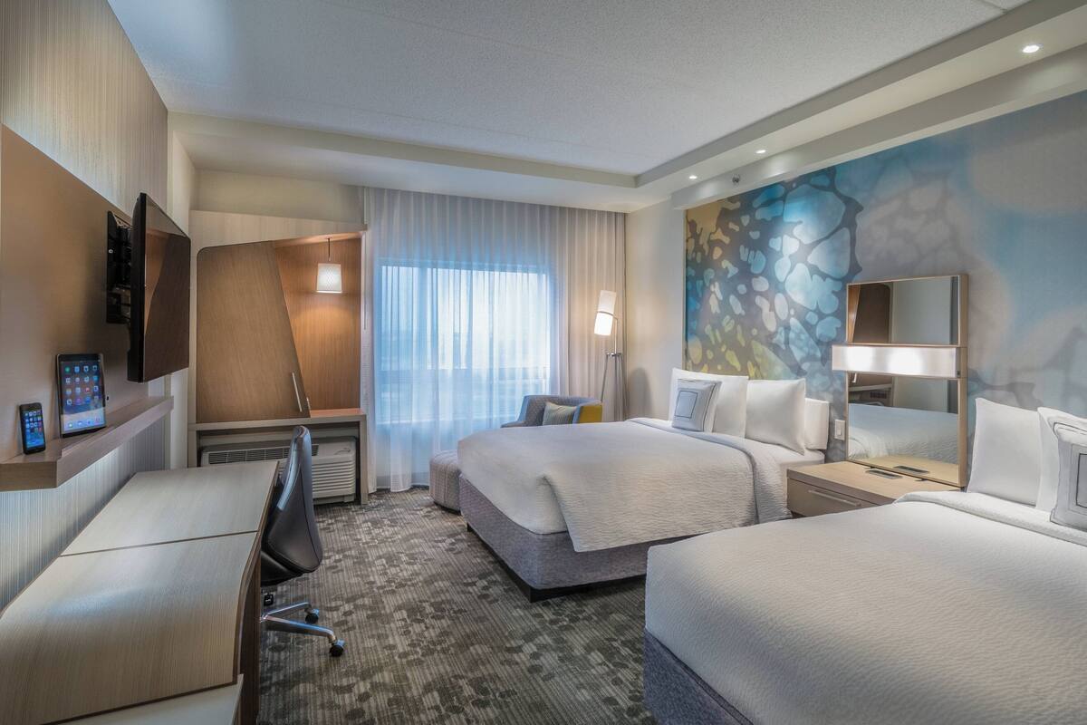 Ein Hotelzimmer mit zwei Betten, einem Schreibtisch, einem Sessel, einem Fernseher und einem großen Fenster mit Vorhängen.