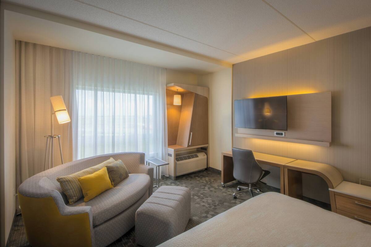 Ein Hotelzimmer mit einer Couch, einem Schreibtisch, einem Sessel, einem Fernseher und einem Fenster mit Vorhängen.