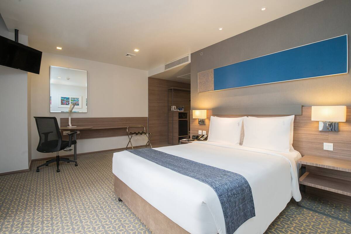 A hotel room with a large bed, a desk, and a chair. There is a TV mounted on the wall and a window with blinds.