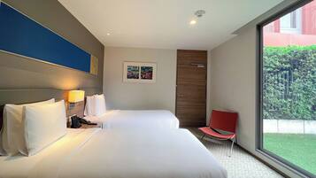 A hotel room with a large bed, bedside tables, a red chair, a window with a view of greenery, and a door.