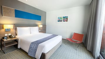 Quarto de hotel com uma cama grande, luminárias de cabeceira, uma cadeira vermelha e um tapete estampado.