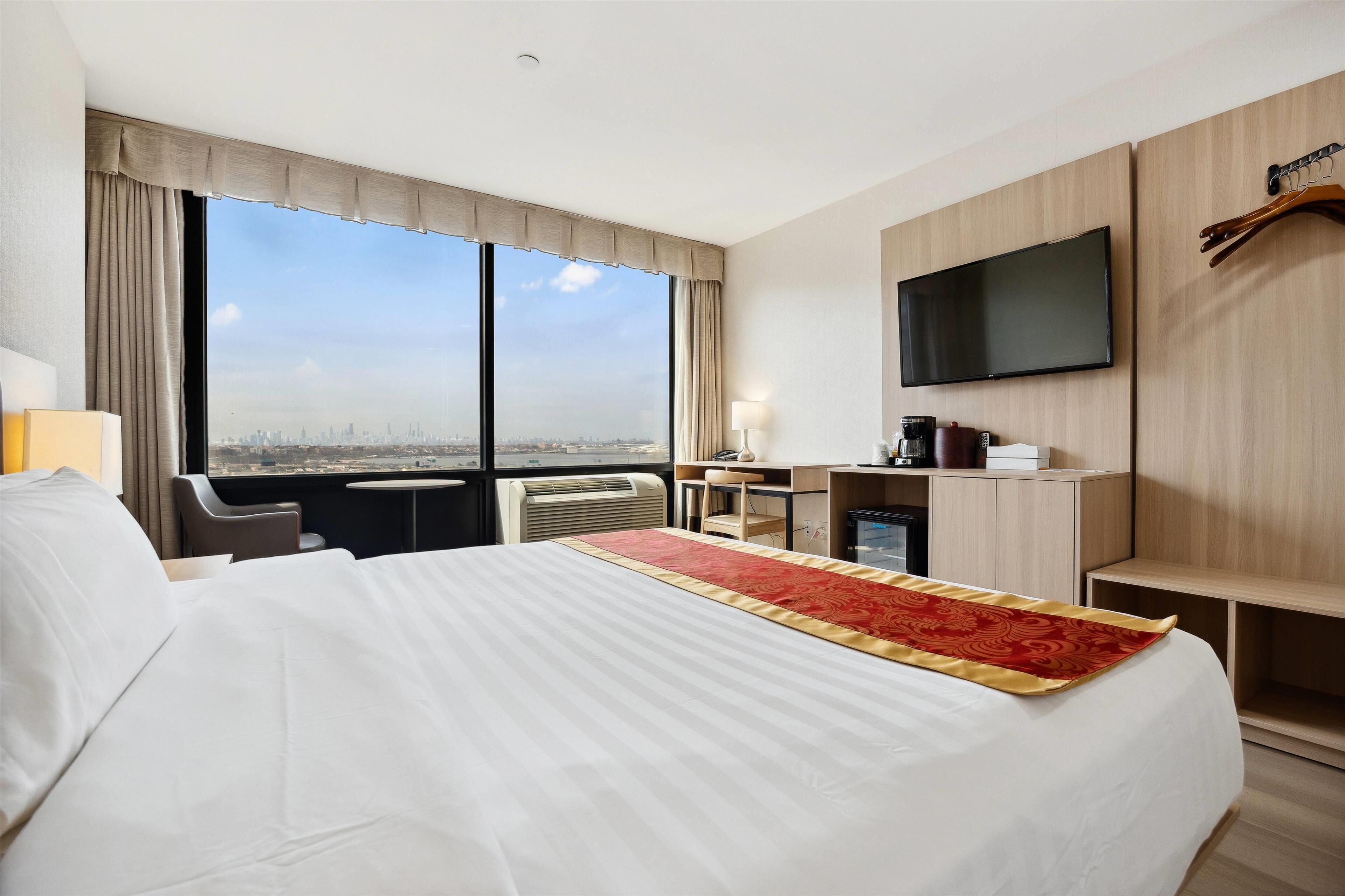 A hotel room with a large bed, a desk, a chair, a TV, and a window with a city view.