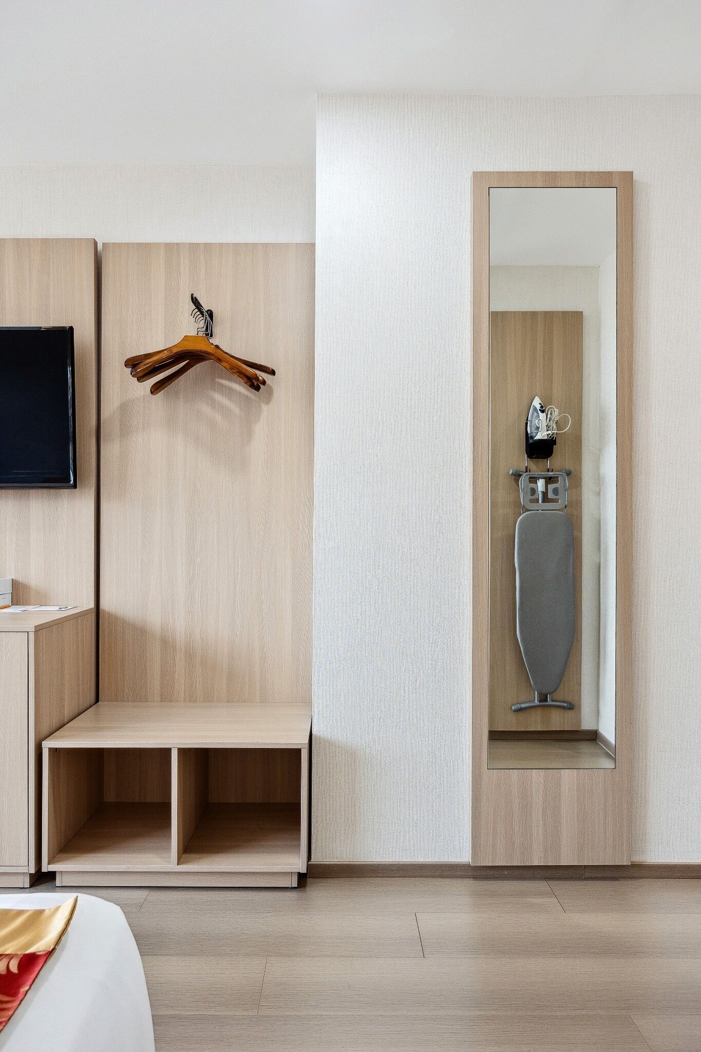 A hotel room with a wooden wardrobe, a mirror, a TV mounted on the wall, and a bench.