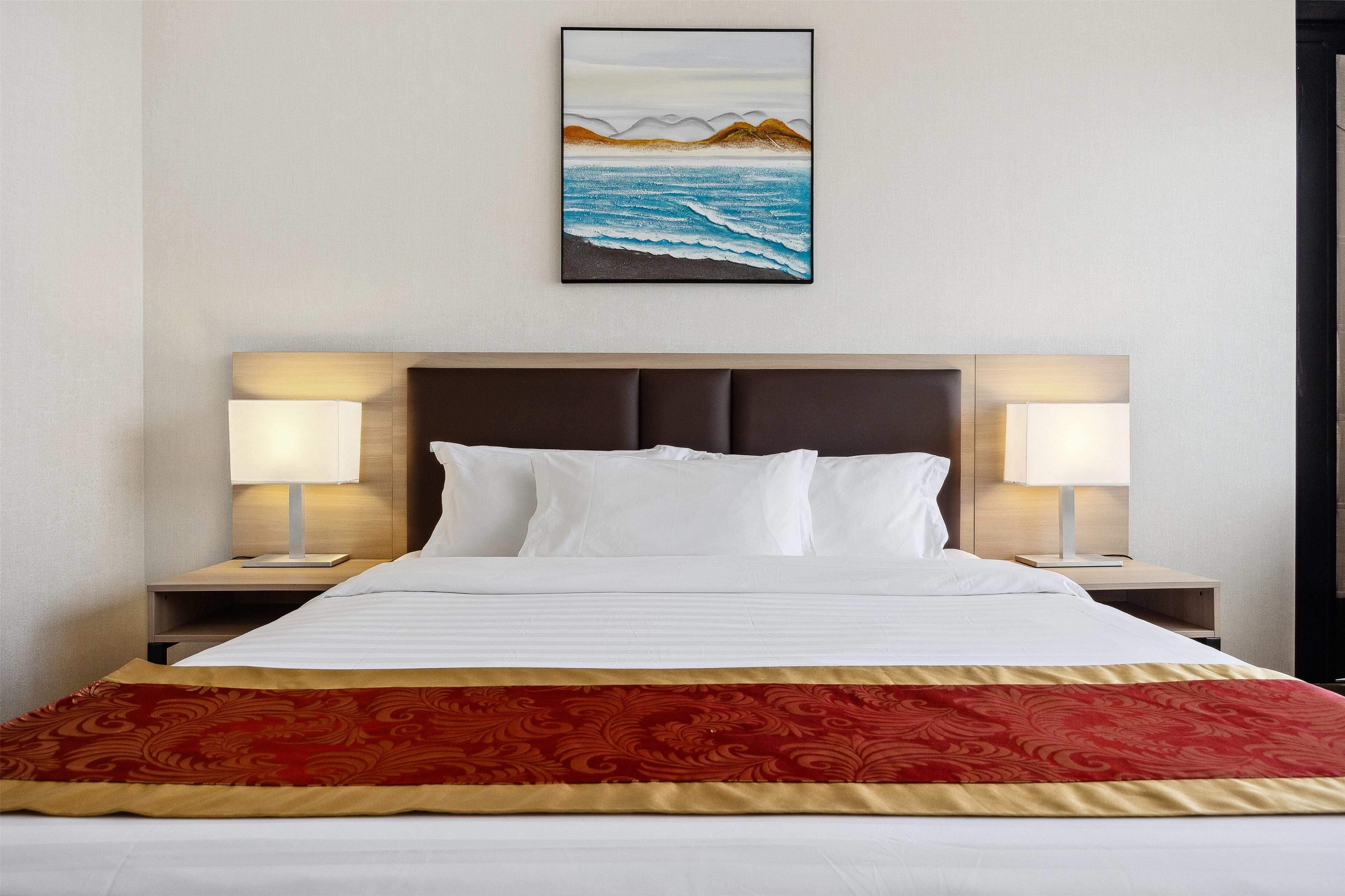 A hotel room with a large bed, two bedside tables with lamps, a headboard, and a painting of a beach scene on the wall.