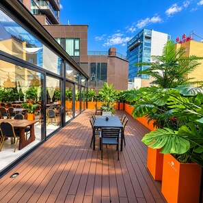An outdoor patio area with tables and chairs, surrounded by potted plants and modern buildings.
