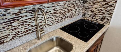 A kitchen with a stainless steel sink, a black stovetop, and a backsplash made of small, rectangular tiles in various shades of brown and beige.