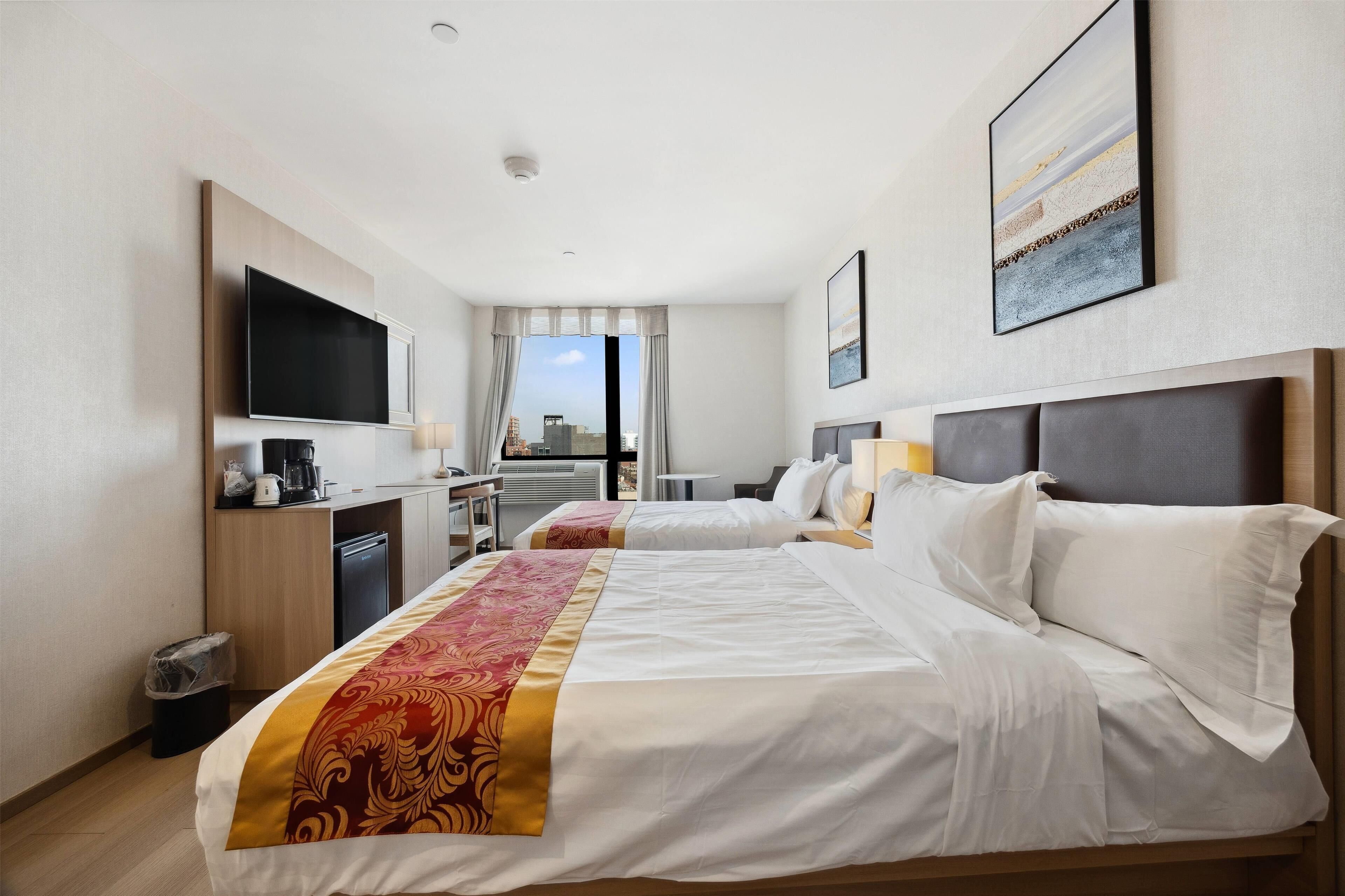 A hotel room with two beds, a TV, a desk, and a view of the city.