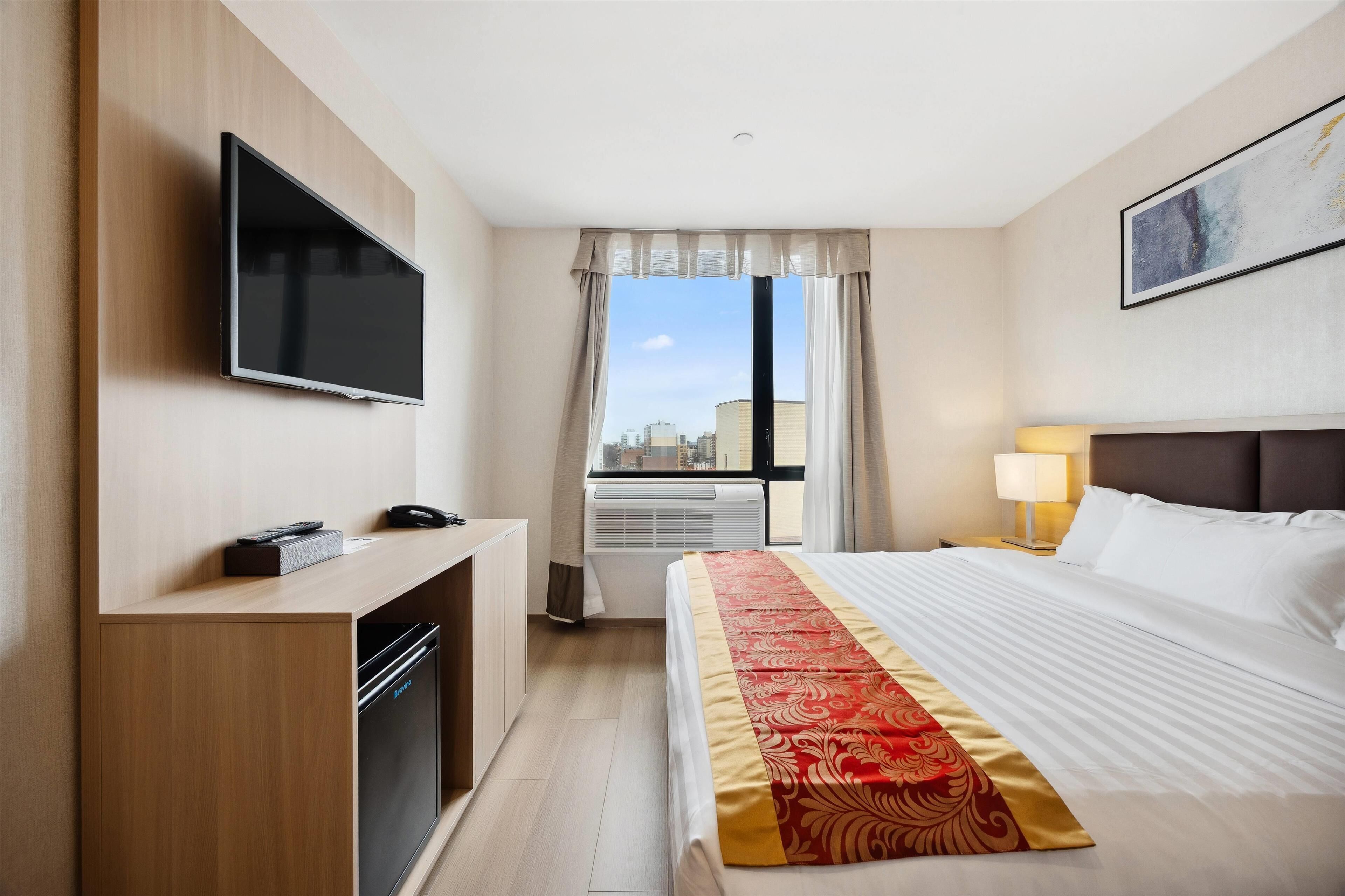 Habitación de hotel con cama, televisor montado en la pared, escritorio con teléfono y ventana con cortinas.