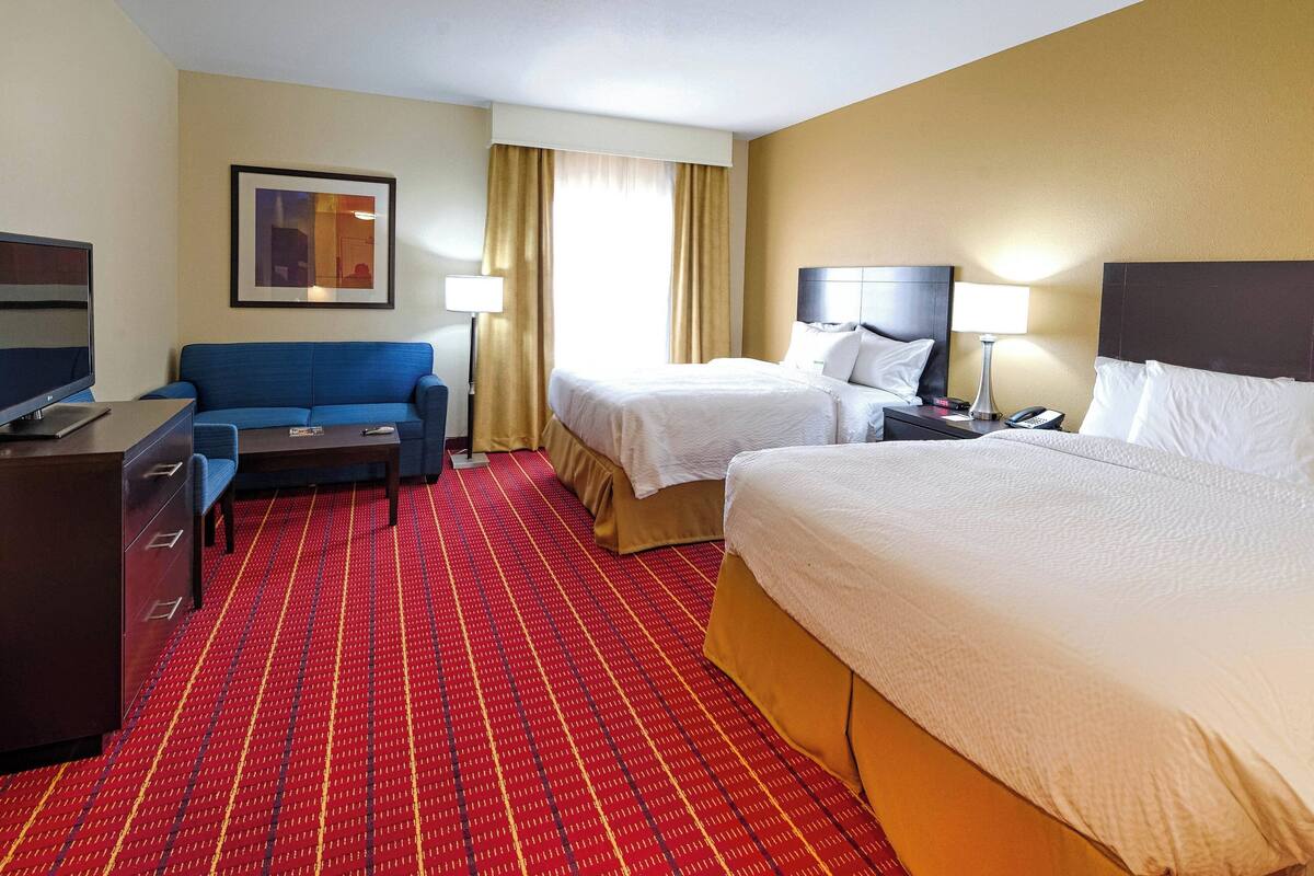 A hotel room with two beds, a desk with a computer, a sofa, and a red patterned carpet.