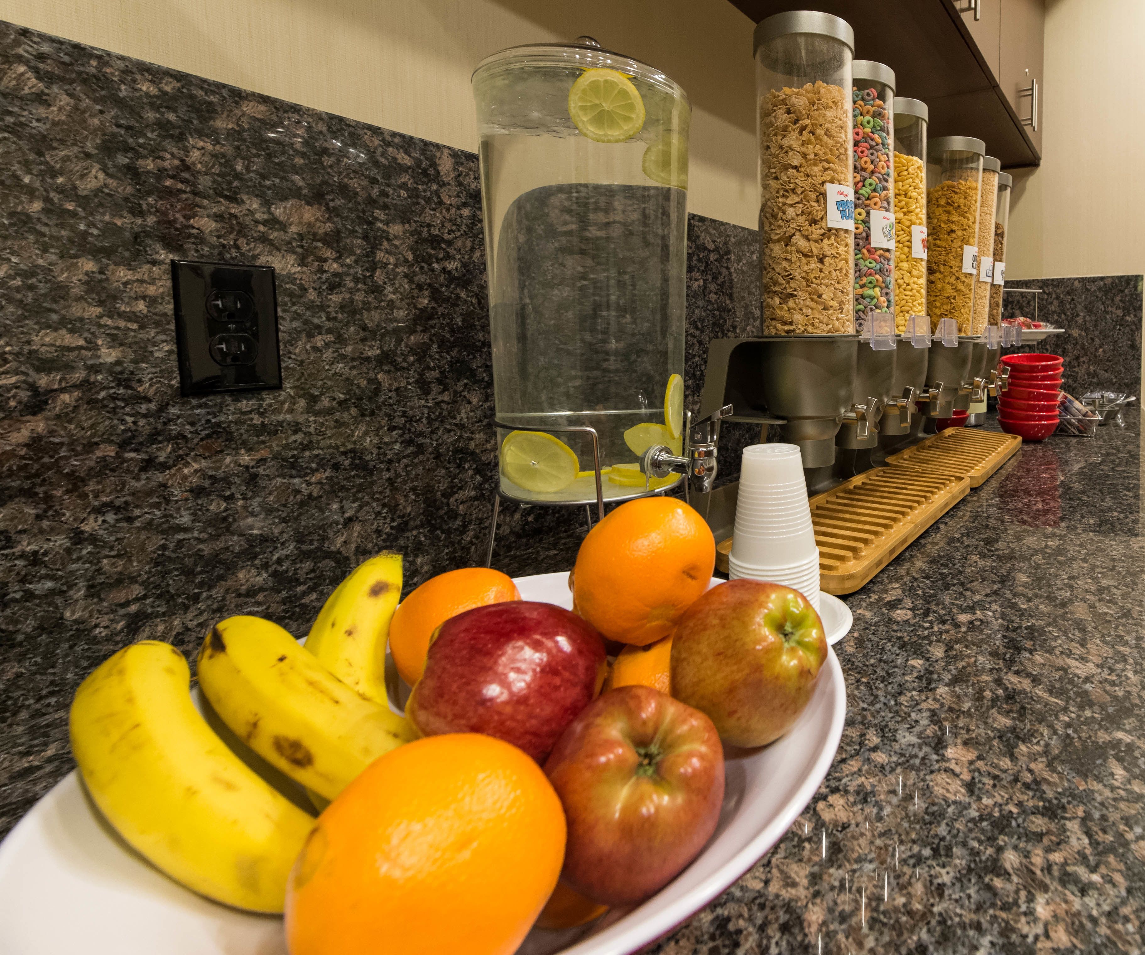 A breakfast bar with fruits, a water dispenser, cereal containers, and a coffee station.