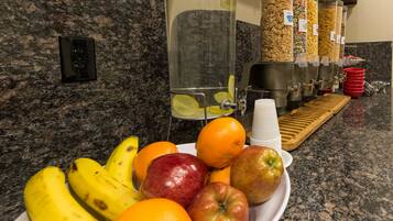 A breakfast bar with fruits, a water dispenser, cereal containers, and a coffee station.