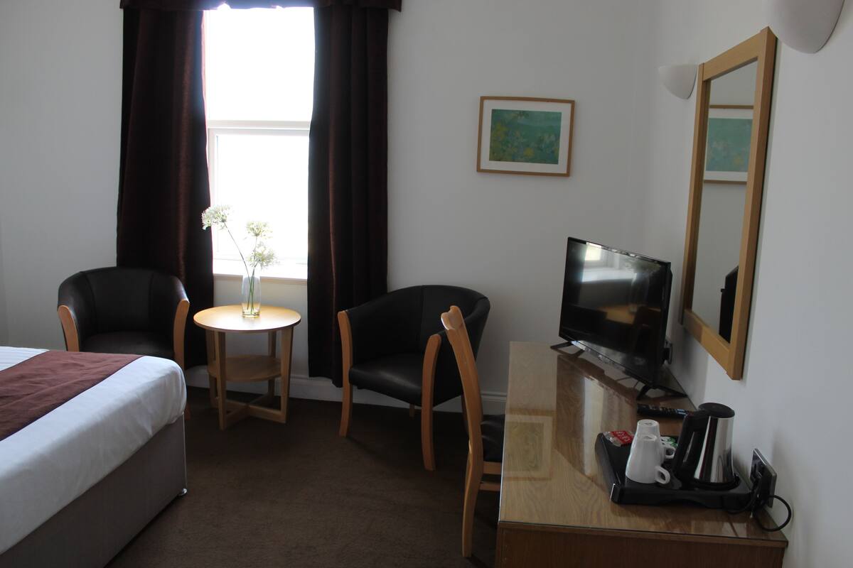 A hotel room with a bed, two chairs, a small table with a vase, a TV, and a coffee maker.