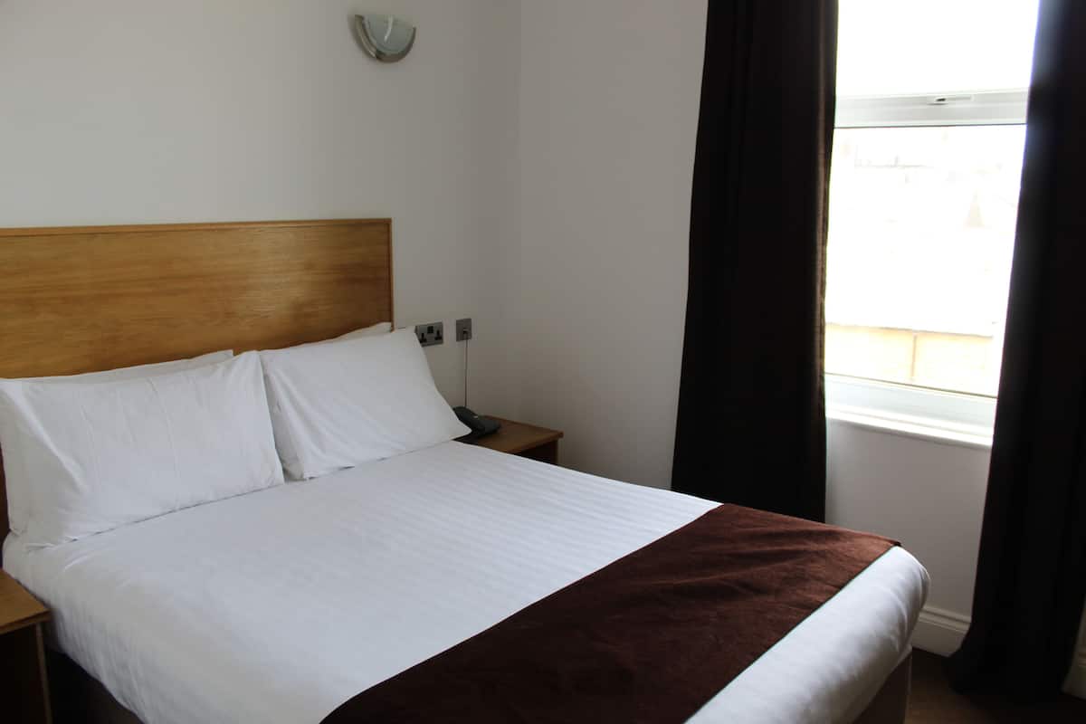 A neatly made bed with white linens and a brown throw blanket, a wooden headboard, a bedside table with a lamp, and a window with curtains.