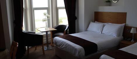 Habitación de hotel con cama, escritorio y silla. Cuenta con una ventana con cortinas, una mesita de noche con lámpara y una pequeña planta en la pared.