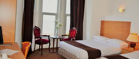 A hotel room with a bed, two chairs, a small table with a vase, a desk with a computer, and a lamp.
