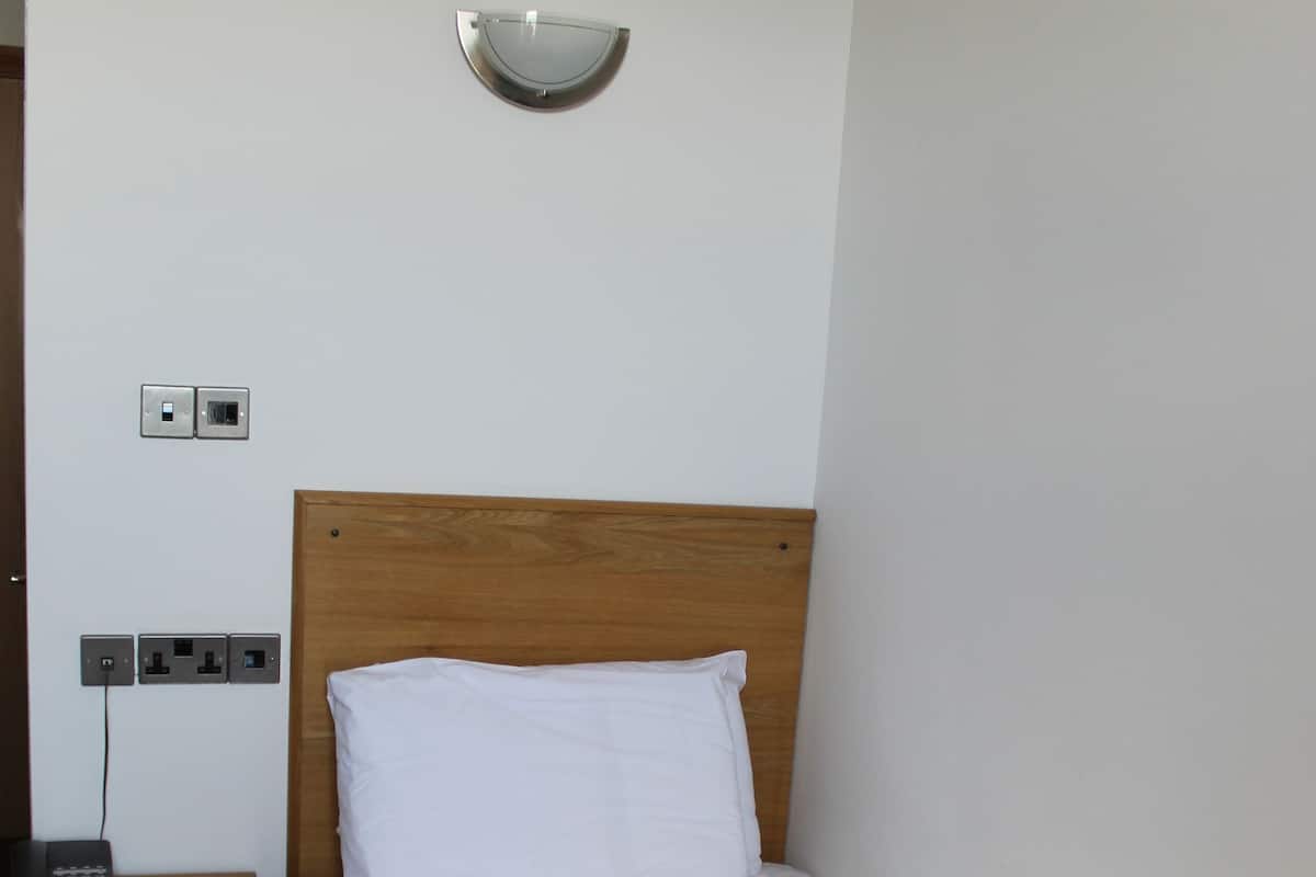 A neatly made bed with a white pillow and a dark brown blanket, a wooden nightstand with a phone, and a wall-mounted light fixture.