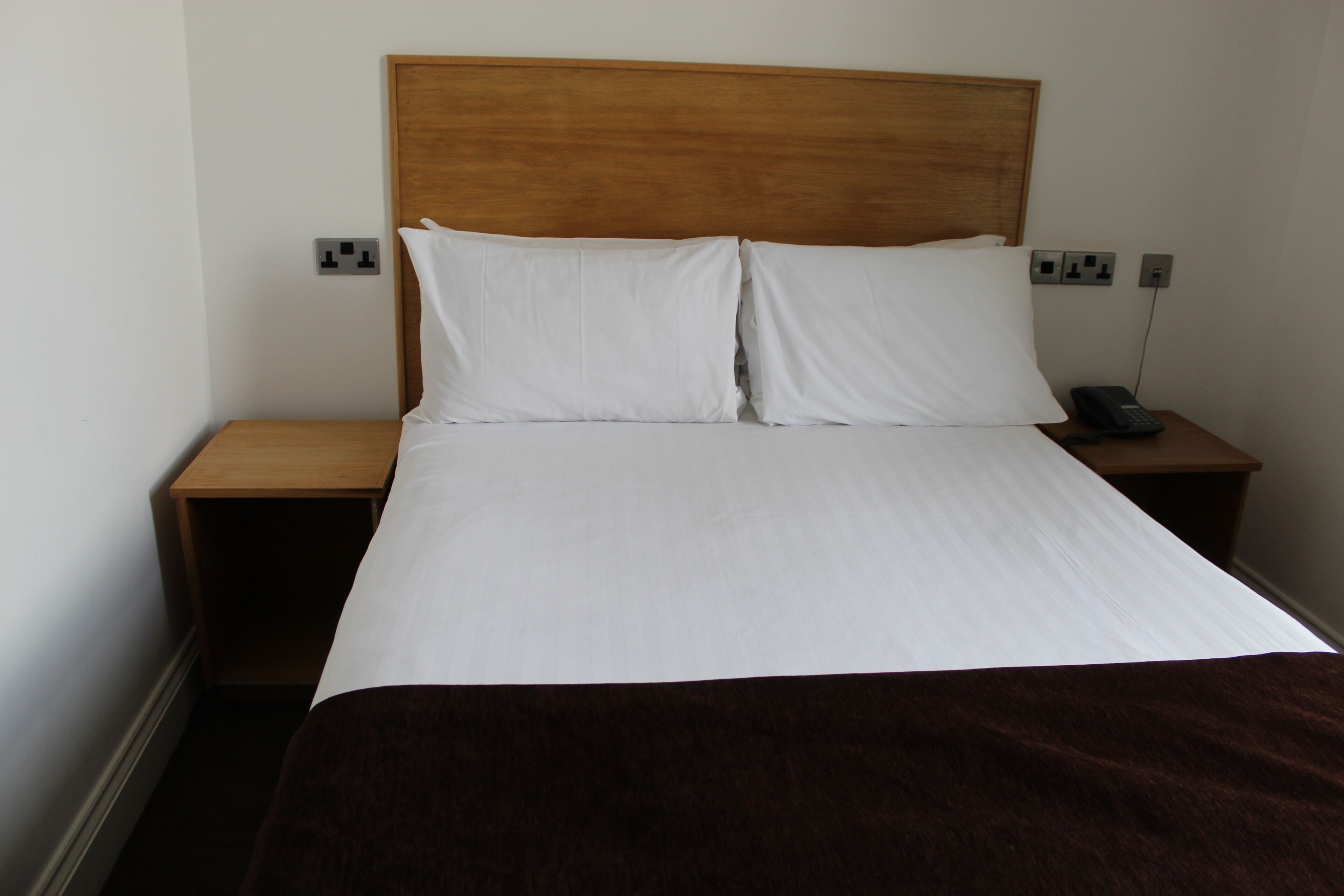 A neatly made bed with white linens and a brown throw blanket, a wooden headboard, and a bedside table with a telephone.