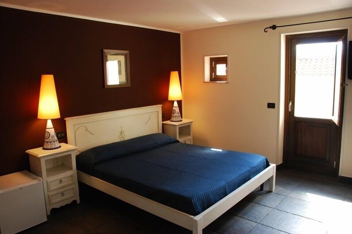 A bedroom with a bed, two bedside tables, two lamps, a small nightstand, and a door.